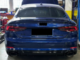 A blue car is parked in an automotive workshop with its hood open. Shelves, tools, and equipment are visible around it. Text on the car reads 