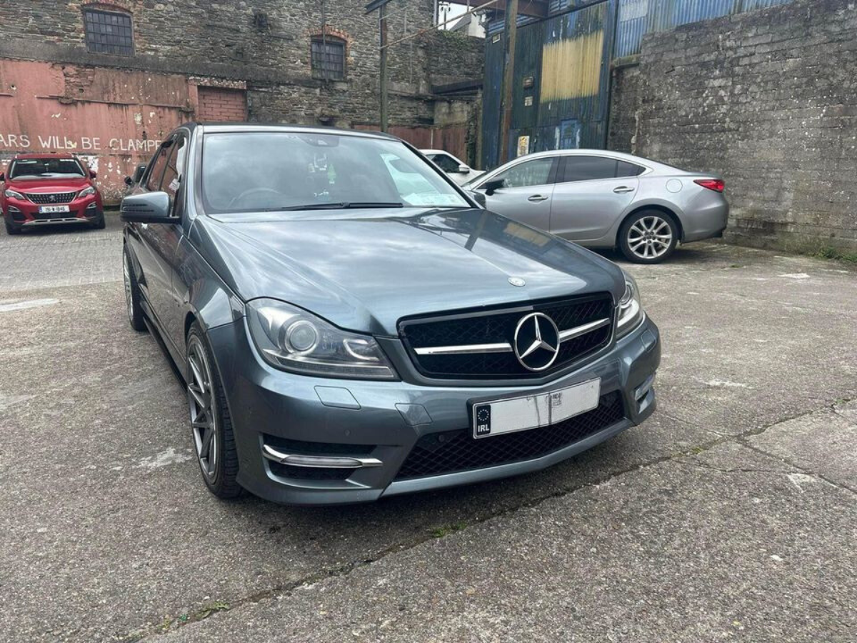 A silver Mercedes-Benz is parked in an industrial yard next to two other cars. The surrounding environment features stone walls and a metal structure with a sign: "CARS WILL BE CLAMPED."