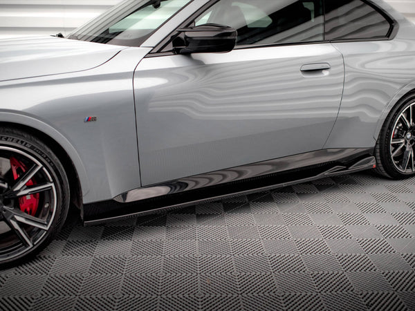 A sleek, silver car is parked on a textured gray floor. It features red brake calipers, sporty black wheels, and a side emblem with colored stripes.