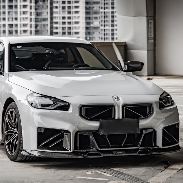 A white, sporty car is parked in a modern urban setting, featuring a bold front grille and sleek design. High-rise buildings are visible in the background.