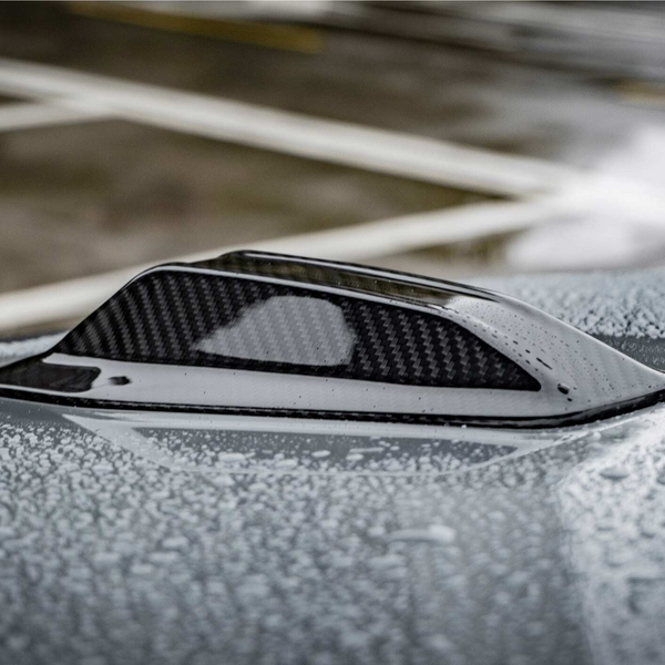 A carbon-fiber shark fin antenna rests on a car roof, covered with water droplets, in a parking lot with painted lines visible in the background.