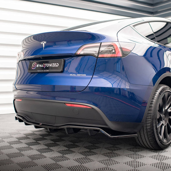 A blue Tesla Model Y with "Dual Motor" label is parked, showing its sleek rear against a patterned floor. The license plate reads "#MAXTONIZED TESLA.COM".