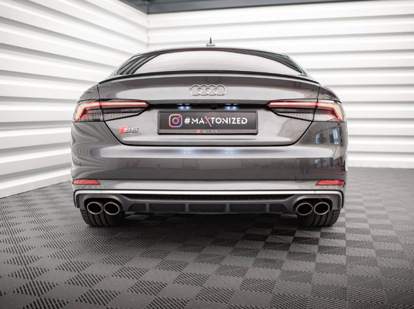 A gray Audi S5 is parked in a brightly lit showroom with white striped walls and a textured floor. The license plate reads "#MAXTONIZED" with an Instagram logo.