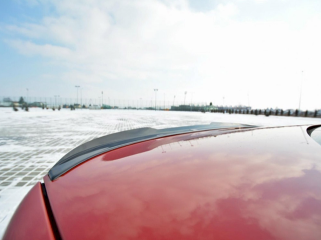 A red car's shiny hood reflects the sky in a wide, snow-covered parking lot with scattered trees and light poles under a cloudy blue sky.