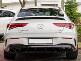 A white Mercedes-Benz CLA 45 S, shown from the rear, displays AMG and model badges. It's parked on a brick surface with trees and a building in the background.