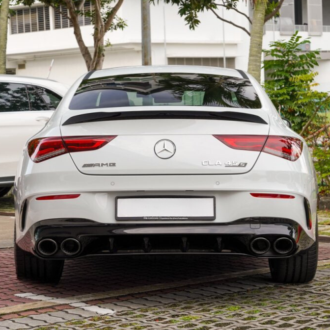 A white Mercedes-Benz CLA 45 S, shown from the rear, displays AMG and model badges. It's parked on a brick surface with trees and a building in the background.