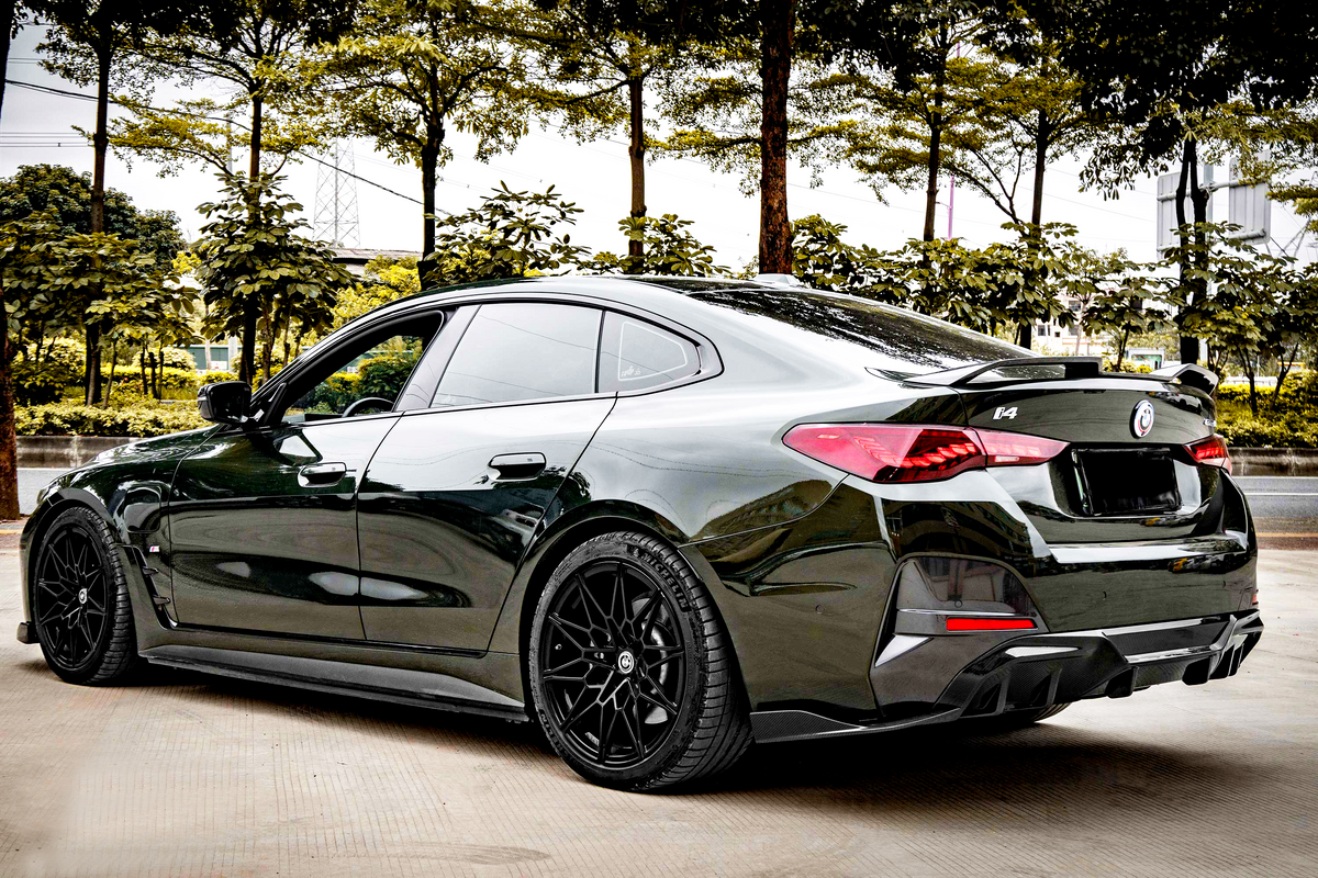 A sleek, dark-colored car is parked on a concrete surface. It features prominent, stylized tail lights and black rims. Trees and shrubs provide a backdrop, suggesting an outdoor setting.