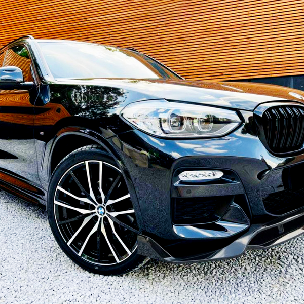 A sleek black car with stylish alloy wheels parked on gravel, positioned in front of a modern wooden slat wall.