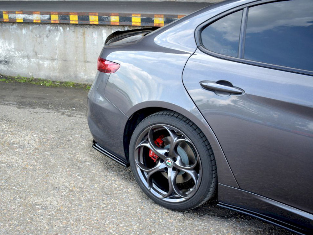 A gray car is parked, showing its rear wheel with red brake calipers. The setting is an outdoor paved area adjacent to a concrete barrier with black and yellow markings.