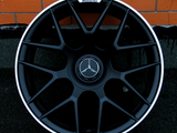 A black alloy wheel with a Mercedes-Benz logo at the center rests against a textured orange brick wall. The top of the wheel features the text 