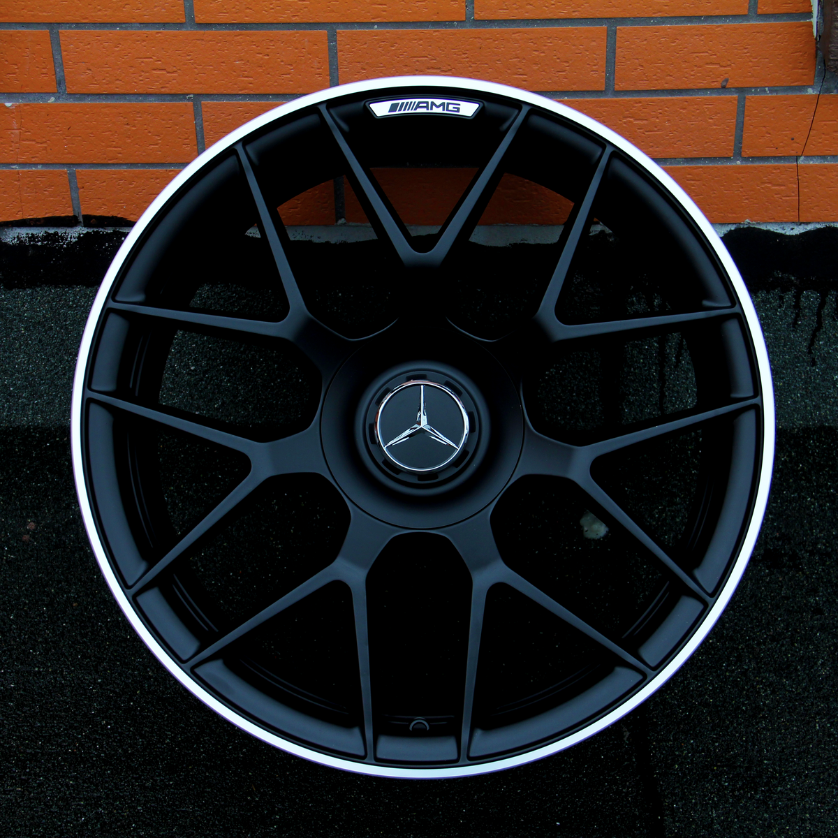 A black alloy wheel with a Mercedes-Benz logo at the center rests against a textured orange brick wall. The top of the wheel features the text "AMG."