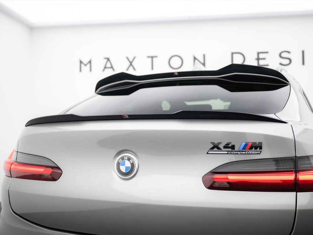 Silver BMW car viewed from the rear, featuring "X4 M Competition" badge. In a showroom setting with "MAXTON DESIGN" on the wall, taillights are illuminated.