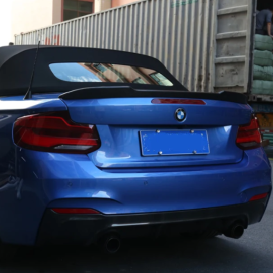 A blue BMW convertible is parked near a large shipping container, with its soft top closed. The surroundings include industrial buildings and stacked goods. The license plate is obscured.