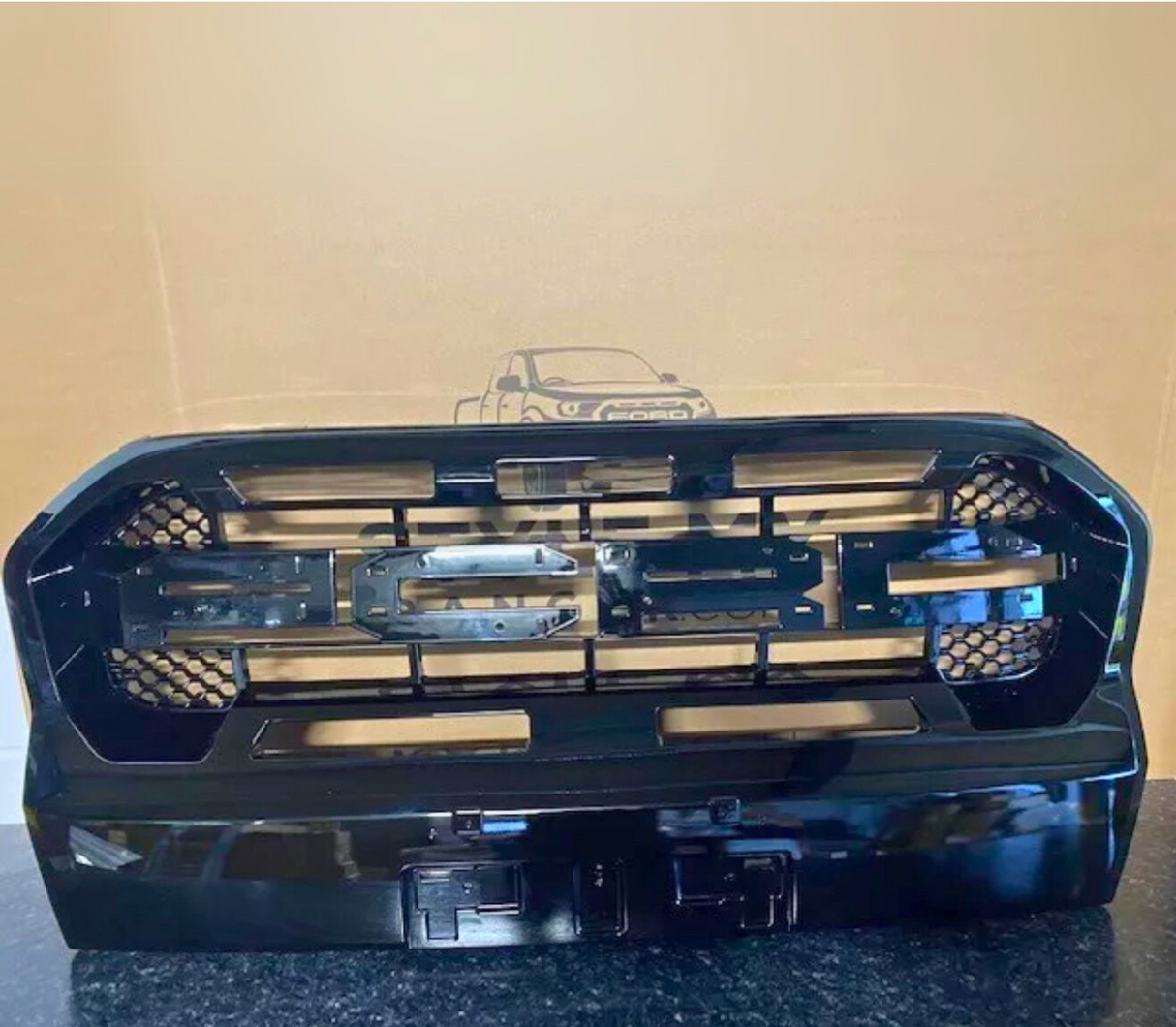 A black car grille, featuring horizontal slats and a honeycomb pattern, rests on a dark surface against a cardboard backdrop with an outline drawing of a Ford vehicle.