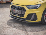 A yellow car's front view, featuring an Audi logo, sporty grille, and headlight on a textured pavement. The license plate reads 