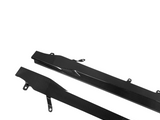 Two black carbon fiber car side skirts, angled with mounting brackets, lie on a white background. Their glossy surface reflects light, highlighting a sleek, textured pattern.