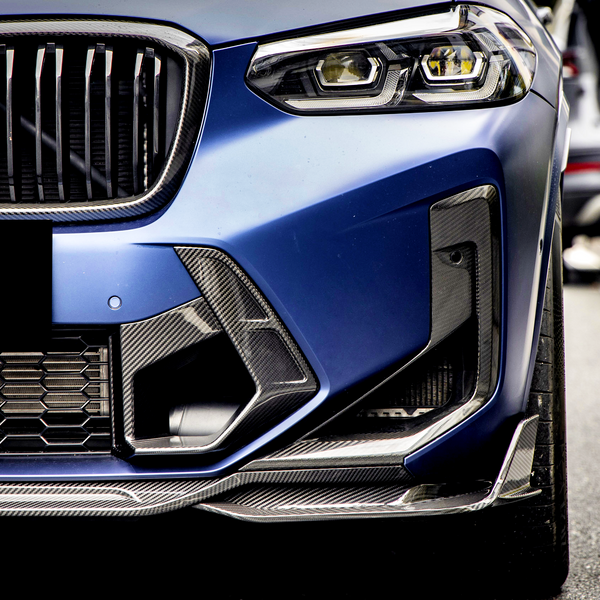 A blue car's front bumper and grille, with a sleek design, is parked on a street lined with cars. The focus is on its headlight and intricate grille pattern.