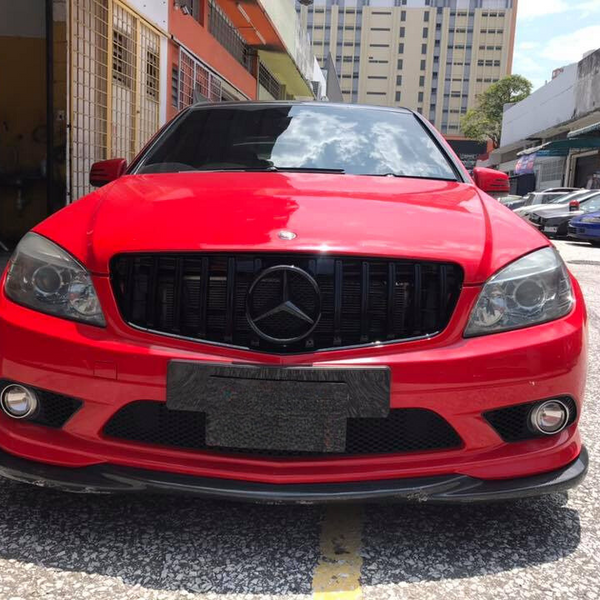 A red Mercedes car is parked on a sunlit street, surrounded by other vehicles and buildings. The front license plate area appears blurred, and the car's grille features the iconic logo.