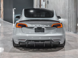 A silver Tesla car is parked in a concrete garage. Its taillights are illuminated, and 