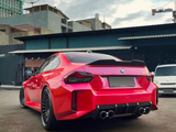 A bright red car is parked, showcasing its sleek design with a sporty spoiler and dual exhausts. It sits in an urban setting with industrial buildings and a cloudy sky.