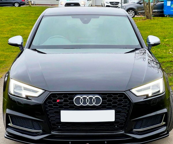 A black Audi S4 is parked on a road, viewed from the front with its headlights on. In the background, other cars and greenery are visible.