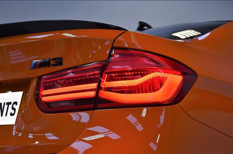 A brightly lit rear taillight on an orange sports car, with the model badge "M3" visible, set against a neutral, indoor background.