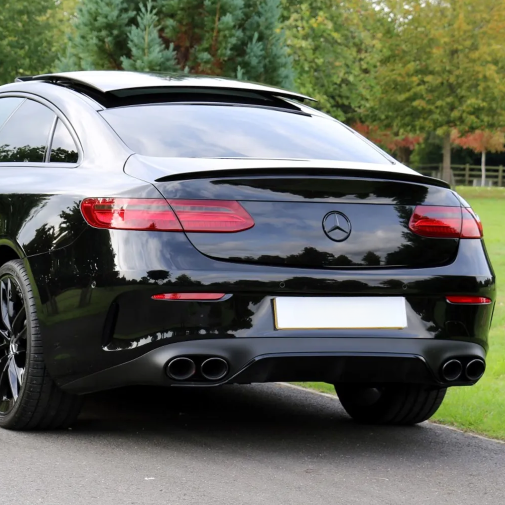 A black car with a sunroof open is parked on a road, surrounded by grass and trees. The rear features dual exhaust pipes and a Mercedes-Benz logo. License plate is blank.