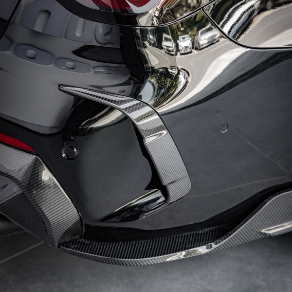 A sleek, glossy car bumper with a carbon fiber fin detail, reflecting nearby buildings, parked on a tiled surface.