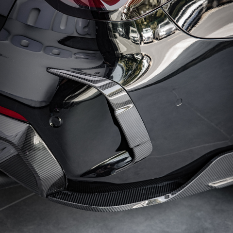 A sleek, glossy car bumper with a carbon fiber fin detail, reflecting nearby buildings, parked on a tiled surface.