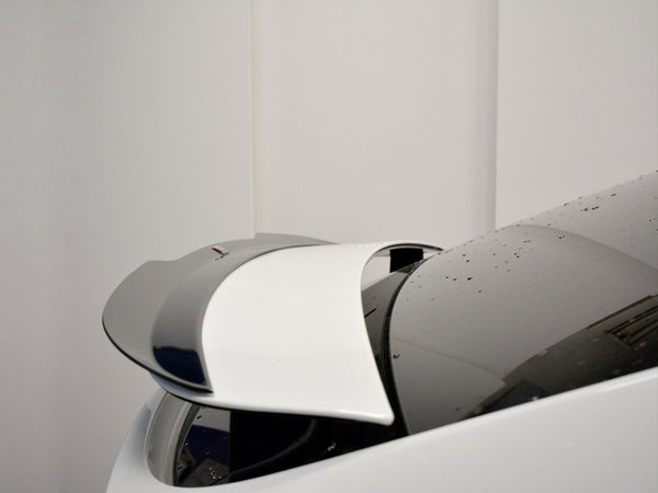 A white car's retractable spoiler is partially deployed, with water droplets on its surface, set against a plain indoor background.