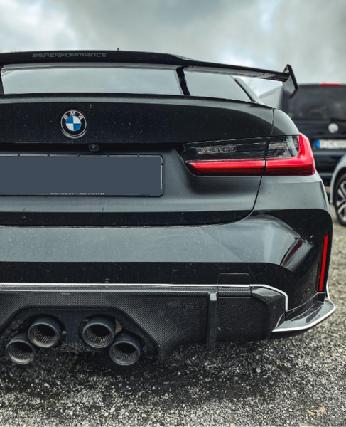 A black BMW car features quad exhaust pipes and a spoiler with "M Performance" text. It is parked on gravel beside another vehicle, under a cloudy sky.