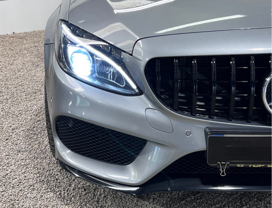A silver car with illuminated left headlight and black grille is parked on gravel. The grille features vertical bars and a circular emblem, while a binder clip secures the front license plate.