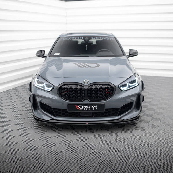 A gray BMW sports car with illuminated headlights is parked in a modern, white-walled showroom. The windshield displays "MAXTON DESIGN." Checkered pattern on the floor.