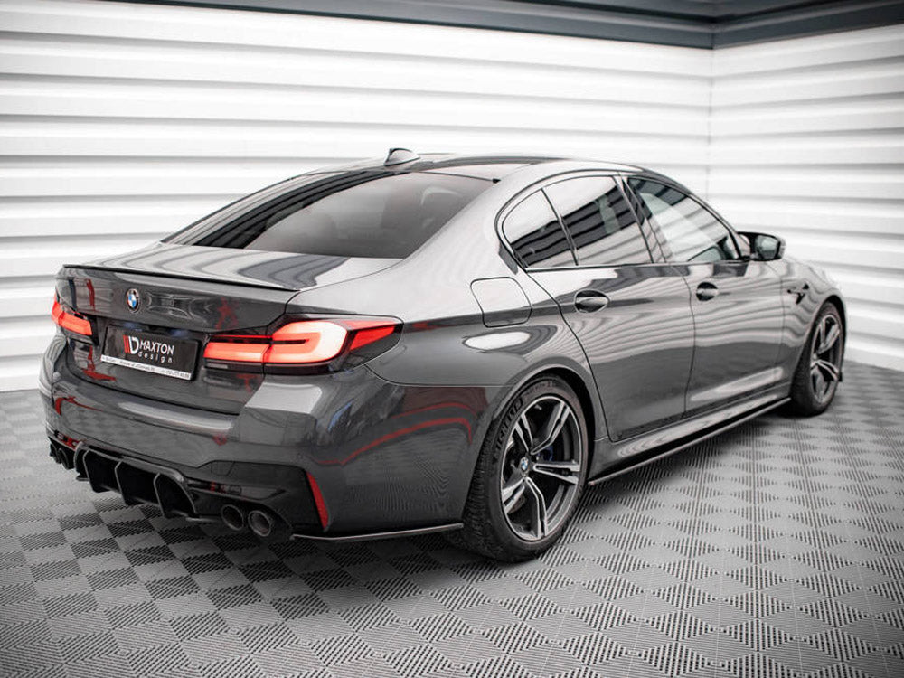 A sleek, gray sedan is parked in a modern, checkered-floor showroom. Its rear features distinctive taillights and dual exhaust pipes. The license plate reads "MAXTON design."