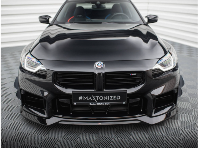 A shiny black BMW car, parked on a patterned surface, features a prominent front grill and headlights. The license plate reads "#MAXTONIZED Dealer BMW M-Cars." The background is a white, corrugated wall.