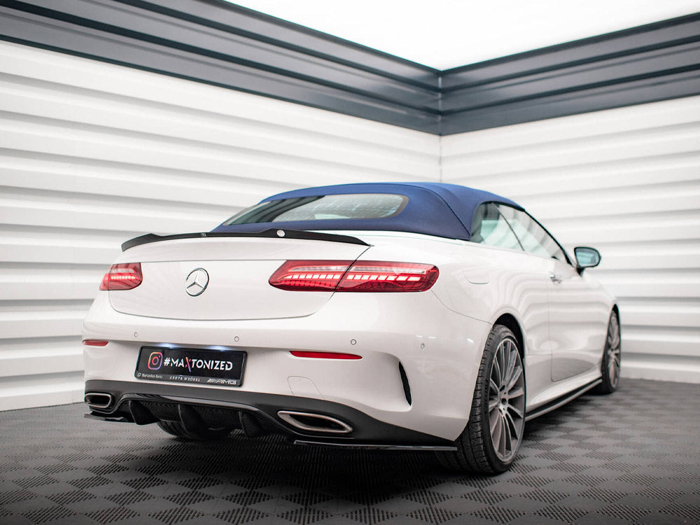 A white convertible car is parked in a modern showroom with white paneled walls. It features a blue roof and visible AMG branding. Text on license plate reads: "#MAXTONIZED."