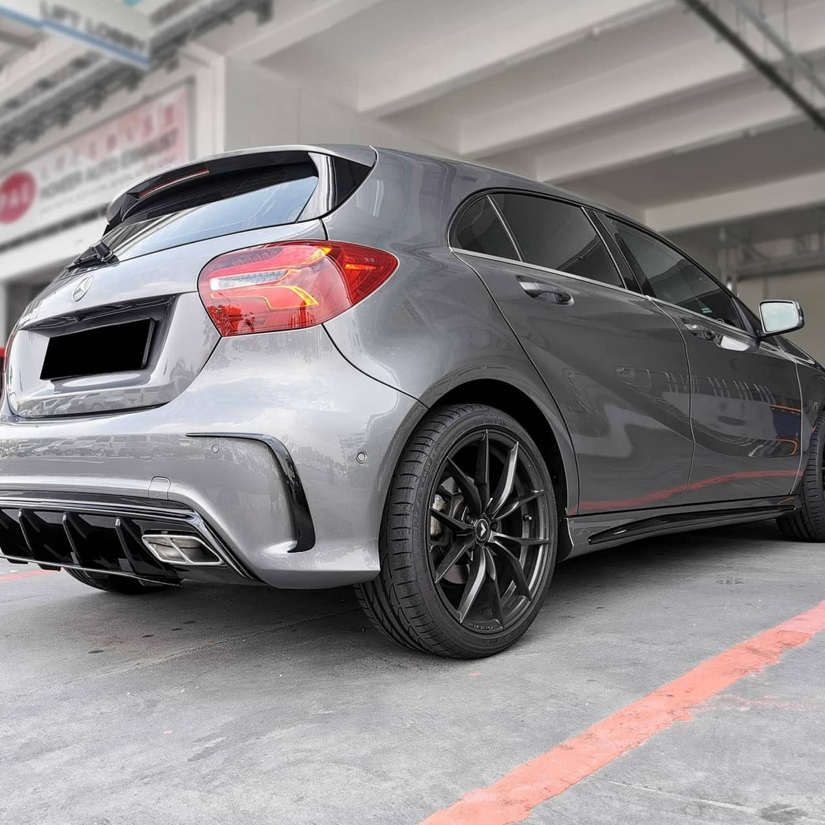 A silver hatchback is parked in a garage-like setting, emphasizing its rear design and black alloy wheels. Surrounding industrial structures create a modern, urban backdrop. No text is visible.