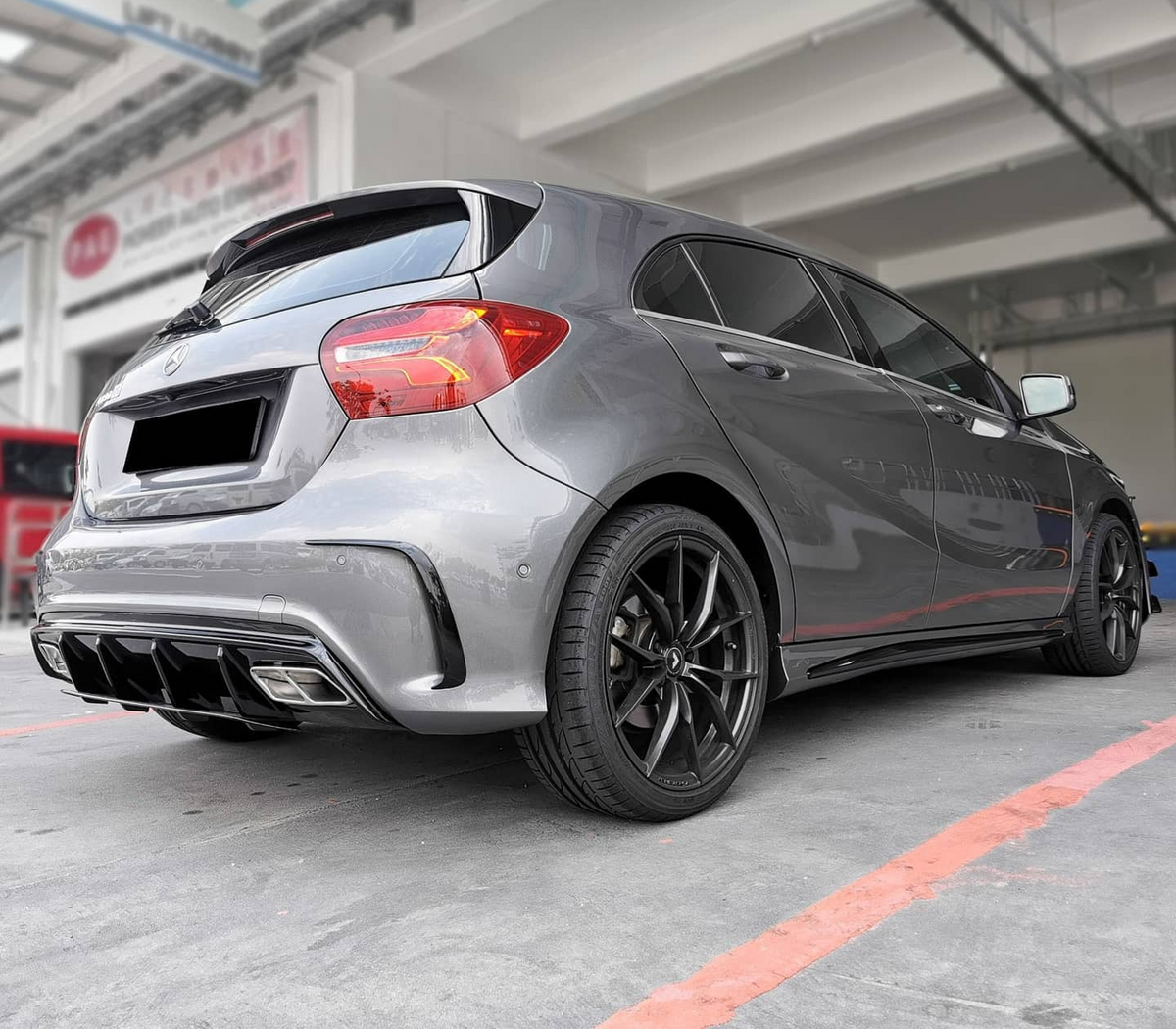 A silver hatchback is parked in a garage-like setting, emphasizing its rear design and black alloy wheels. Surrounding industrial structures create a modern, urban backdrop. No text is visible.