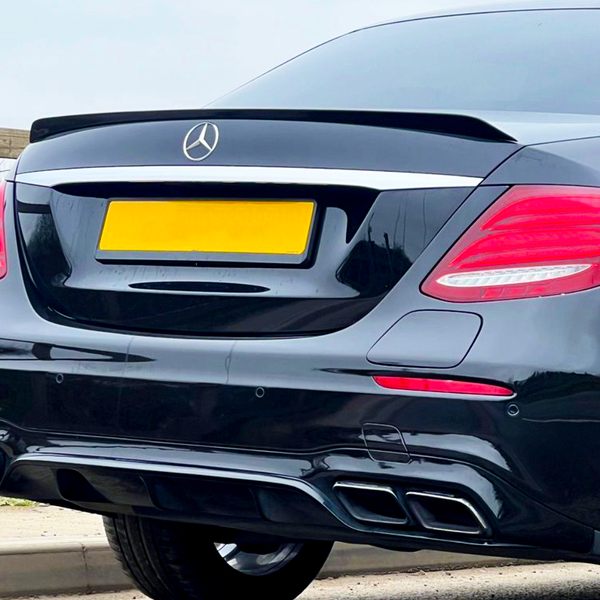 A black car with a visible rear, featuring Mercedes emblem, tinted windows, and dual exhausts, is parked on a road near wooden fencing and greenery. The license plate is yellow.