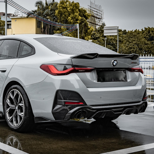 A grey sports car is parked on a wet surface, viewed from the rear, with a visible BMW logo. The background includes trees, a yellow building, and overcast sky.