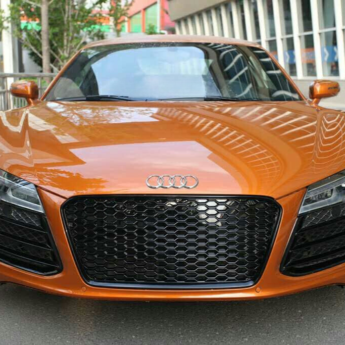 An orange Audi car, parked on a city street, displays a honeycomb grille and Audi emblem, with modern buildings reflecting on its shiny surface.