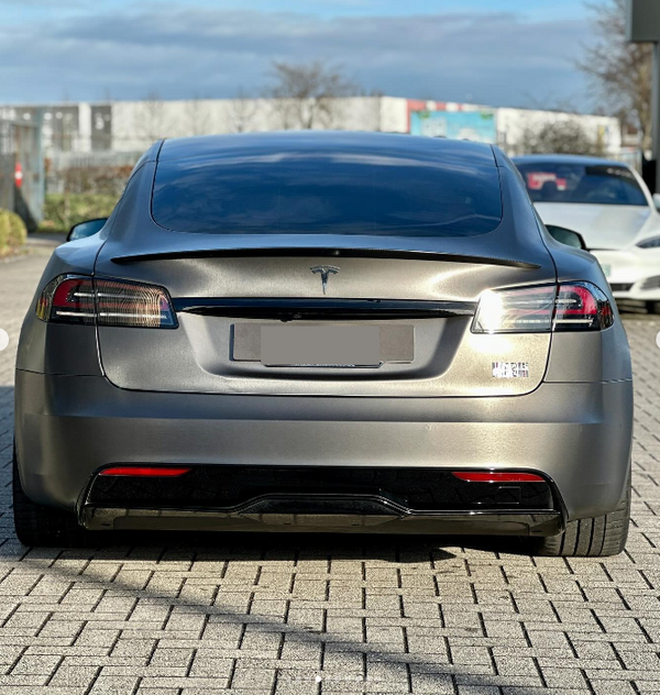 A gray Tesla Model S is parked on a brick driveway, surrounded by other vehicles. The license plate is blurred, and there are trees and buildings in the background.