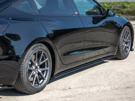 A sleek black car is parked on a sandy path beside a green area. The car's reflective surface mirrors the surrounding trees and sky.