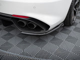 Car exhaust pipes protrude from the rear of a white vehicle, parked on a gray, textured, diamond-patterned surface, with a visible rear tire and partial red reflector.
