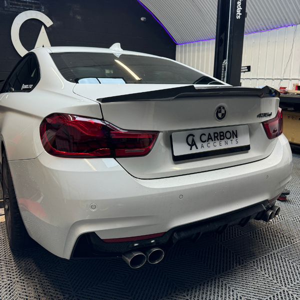 A white BMW 435d featuring a carbon fiber spoiler is parked inside a garage with a sign reading "CARBON ACCENTS." The garage has a dark back wall and a ribbed ceiling.