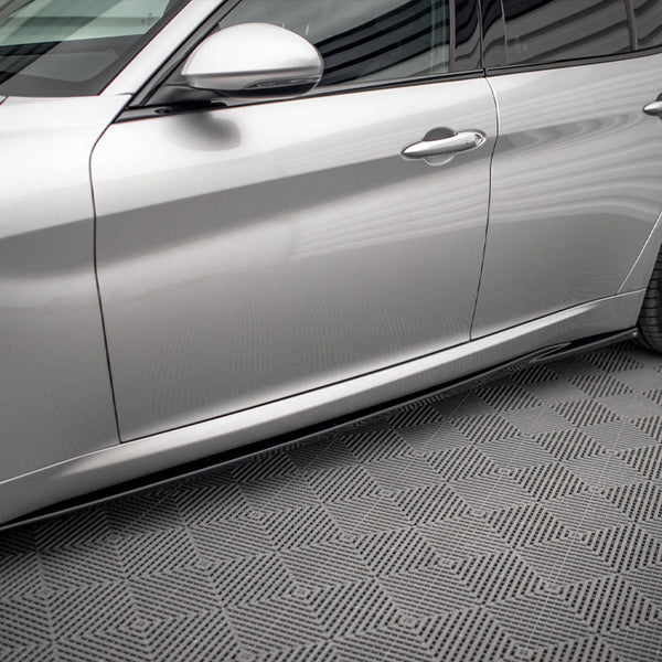 A silver car with sleek, aerodynamic design is parked on a textured, geometric-patterned floor. The car's side doors and black alloy wheels are visible, reflecting soft lighting.