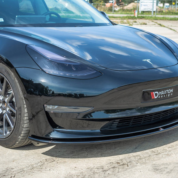 A black sports car is parked on a concrete surface, displaying a "Maxton design" splitter and "Tesla" logo. The environment includes a faint view of grass and fencing in the background.