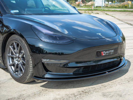 A black sports car is parked on a concrete surface, displaying a "Maxton design" splitter and "Tesla" logo. The environment includes a faint view of grass and fencing in the background.