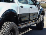 A lifted, black-and-white pickup truck is parked on a road, featuring large off-road tires and side steps with orange reflectors. It’s set against a blurred background.