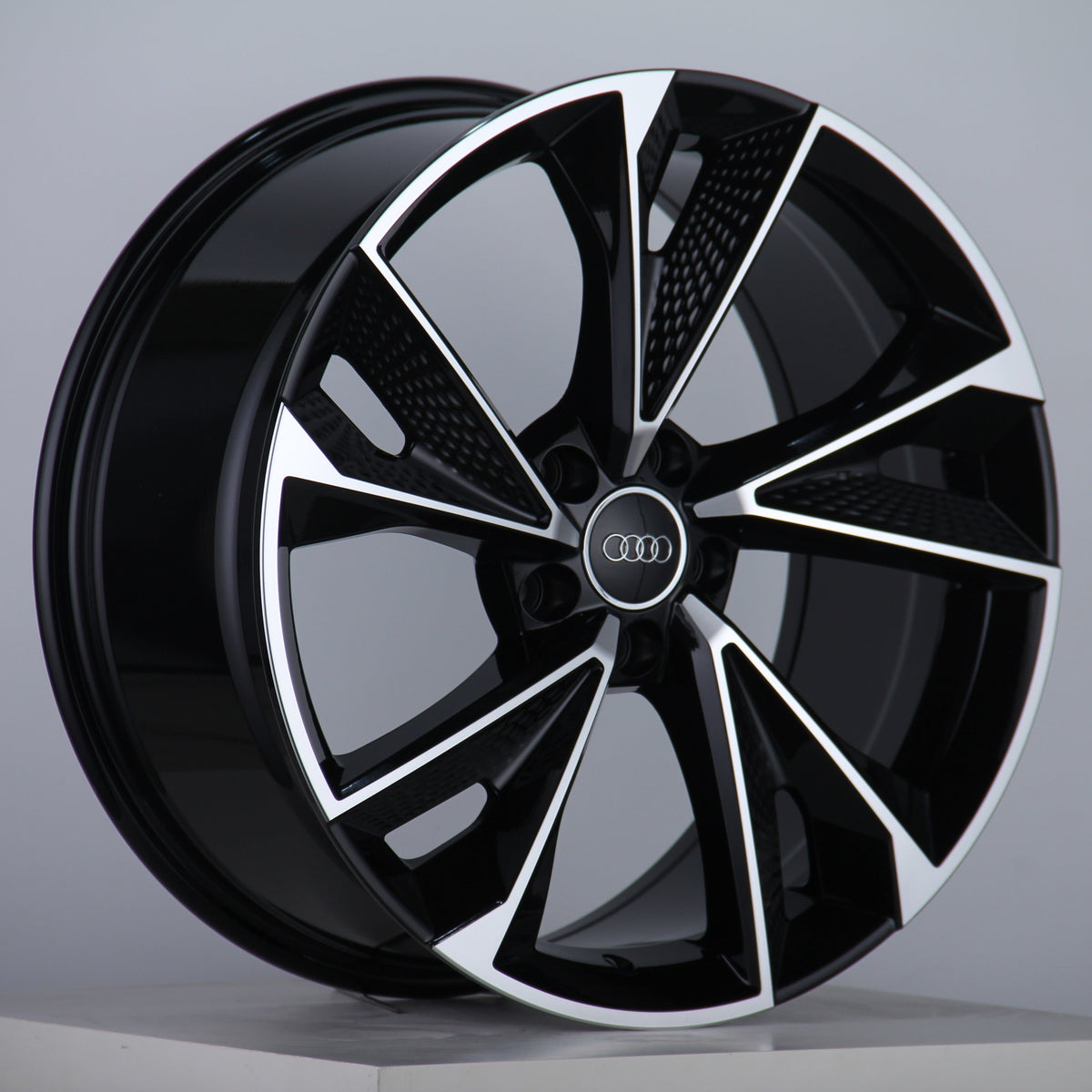 A glossy, black alloy wheel features silver accents and the Audi logo at the center, displayed on a neutral gray background with a studio setting.